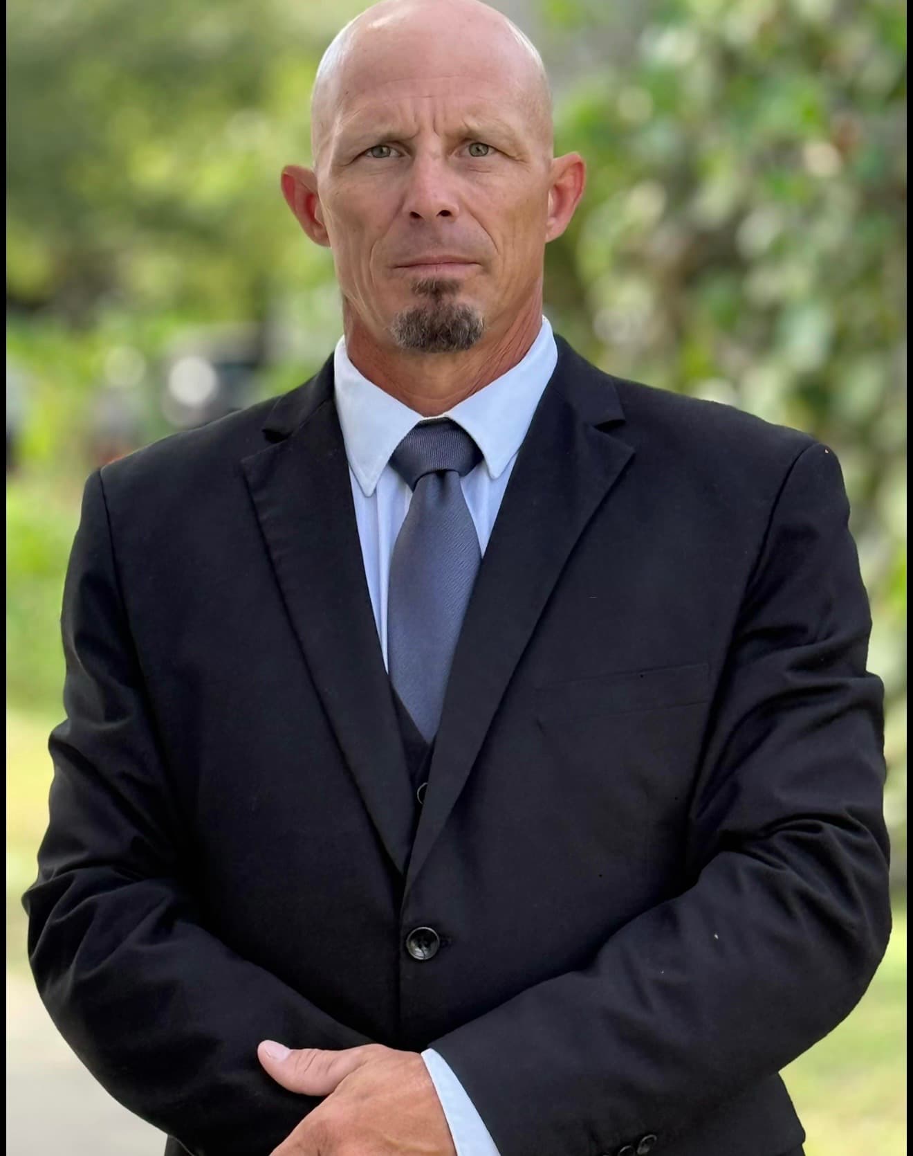 Serious bald man with a goatee wearing a black suit, white shirt, and grey tie.