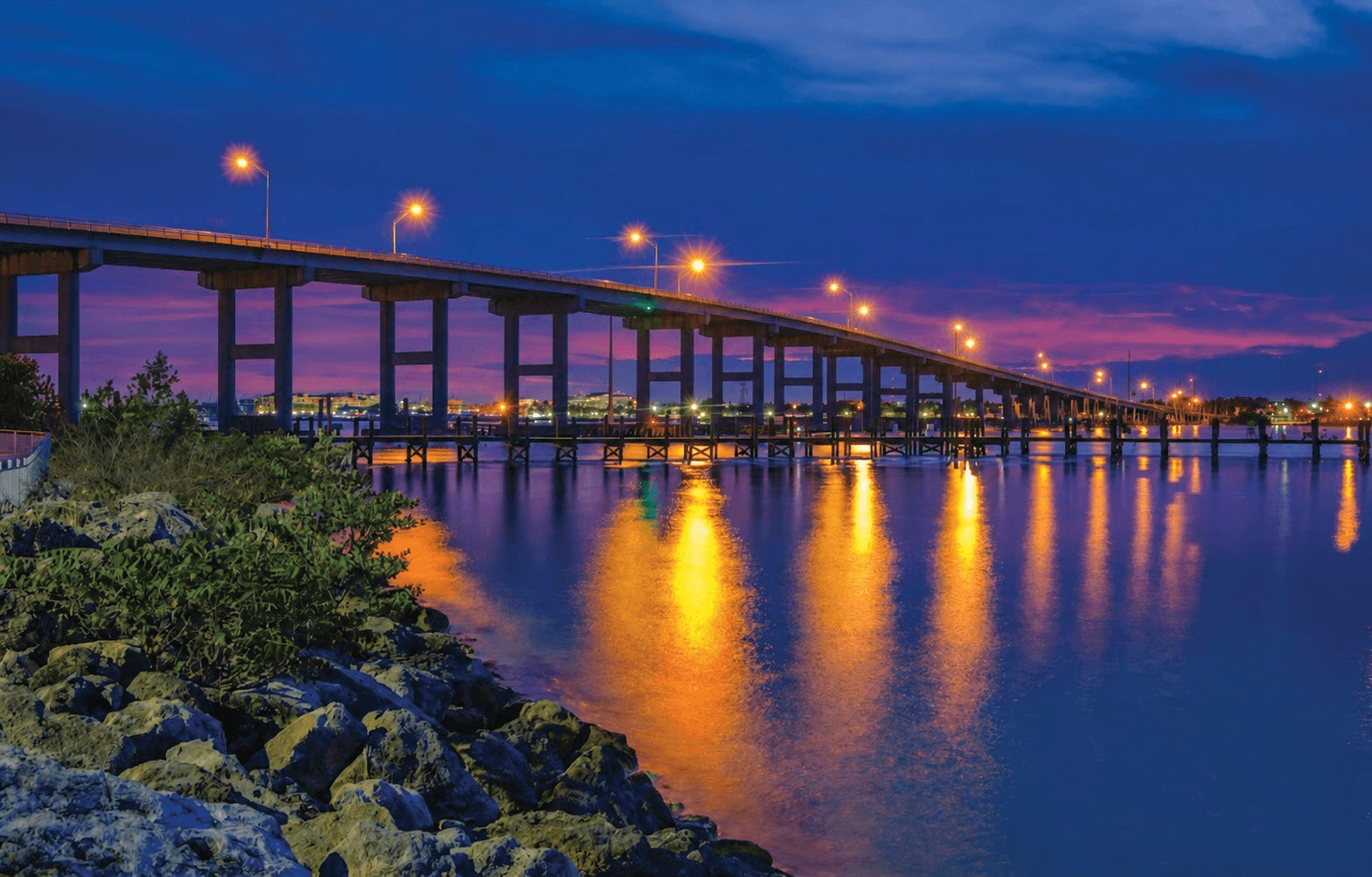 Illuminated bridge over water reflecting golden streetlights under a vibrant purple and blue sunset.