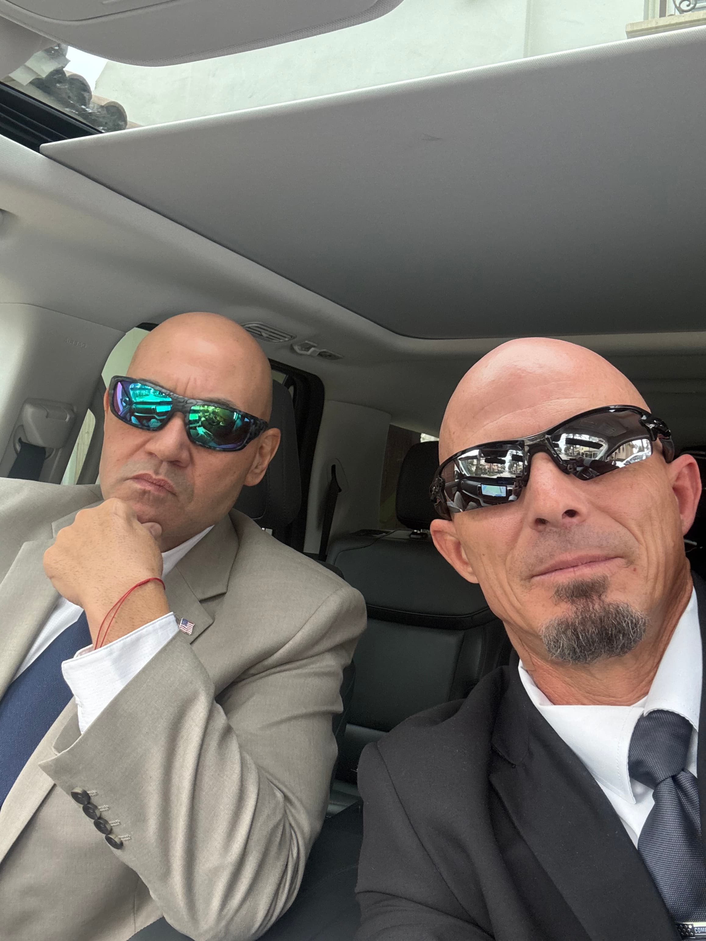 Two bald men in suits and sunglasses sitting inside a car with a sunroof.