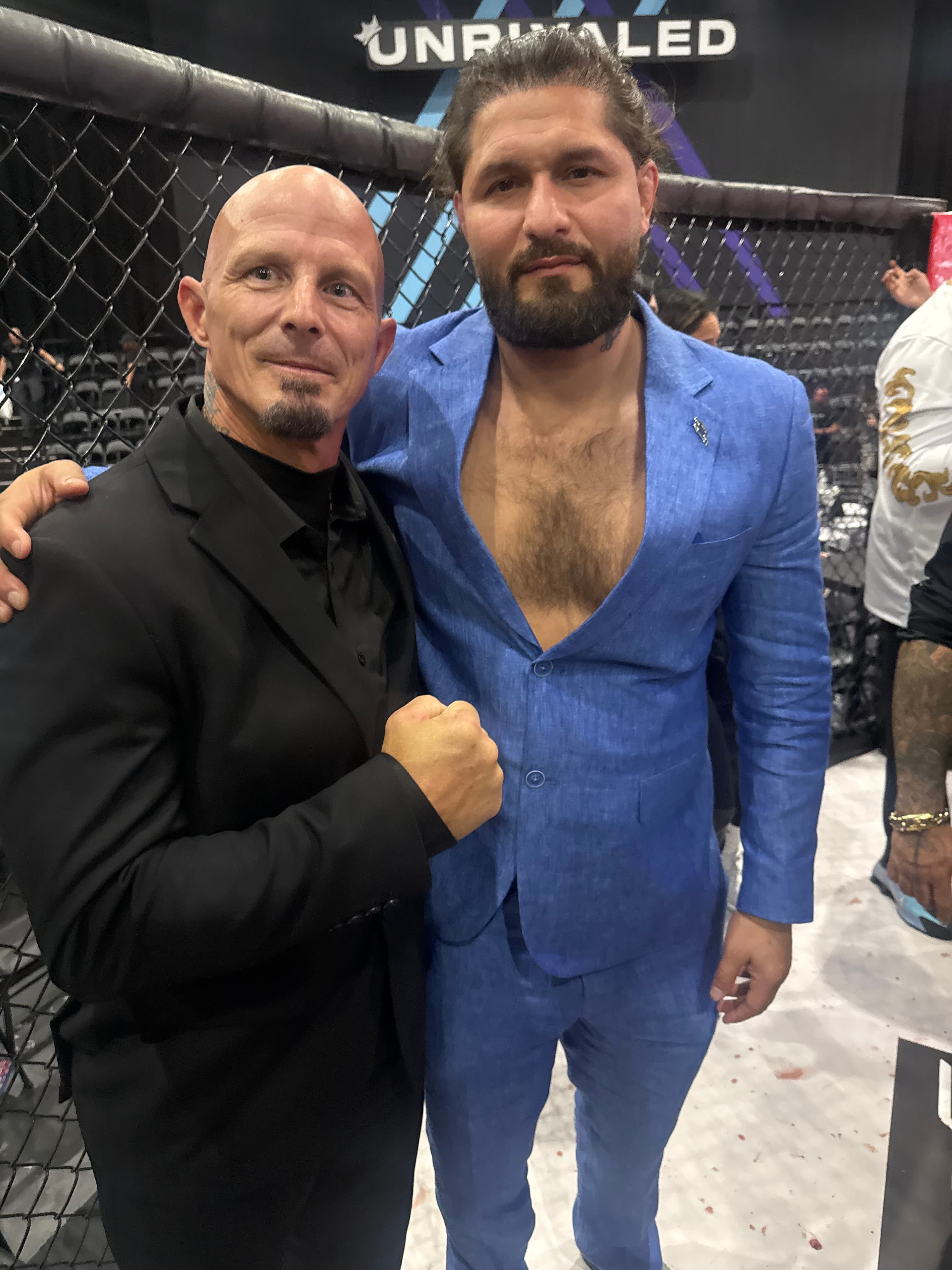 Jorge Masvidal in a blue suit and a bald man pose by a cage.